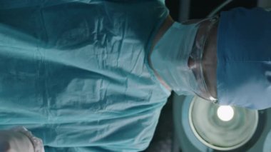 Dikey orta boy, iş kıyafetleri içindeki erkek doktorun ameliyat odasında ciddi bir ameliyat yaparken aşağıya bakması.