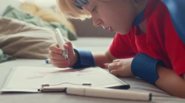 Kafkasyalı çocuk göğsünde kırmızı ve mavi süper kahraman kostümü giyip evde oynarken boya kalemleriyle boyama sayfasına resim çiziyor.