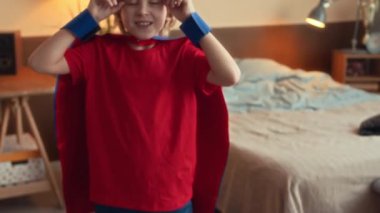 Süper kahraman kostümü giyen, mavi maske takan ve güçlü pazılarını kameraya gösteren 9 yaşındaki neşeli beyaz çocuğun orta yavaş portresi.