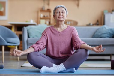 Gri saçlı son sınıf kadınının uzun portresi evde yoga yapmaktan zevk alıyor ve Lotus pozisyonunda gözleri kapalı meditasyon yapıyor.