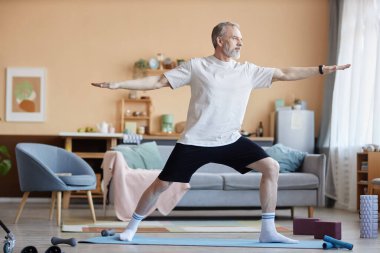 Evdeki fotokopi salonunda yoga yaparken savaşçı pozisyonunda duran güçlü bir kıdemli adamın tam boy yan görüntüsü.