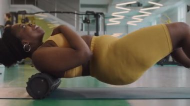 Spor salonunda yağ yakma egzersizi yaparken sırtüstü uzanan neşeli, iri yarı, siyah bir kadının yan görüntüsü.