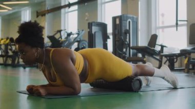 Sarı aktif giysili genç kıvrımlı siyah kadın spor salonunda yoga minderinin üzerinde dururken köpük silindir kullanıyor.