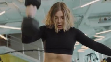 Spor salonunda spor yaparken tek eliyle dambılı kaldıran siyah aktif giysili, kararlı, beyaz bir kadının çekimleri.