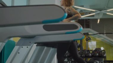 Beyaz tenli, dar etek giyen, spor salonunda koşu bandında koşan genç bir kadının yan görüntüsü.