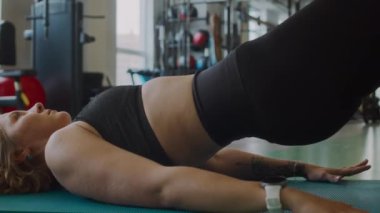Kafkasyalı genç bayan sporcu spor salonunda antrenman yaparken minder üzerinde glute briç oynuyor.