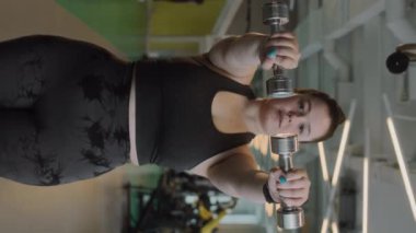 Dikey olarak, spor salonunda dambıllarla egzersiz yapan, kıvrımlı, genç, beyaz bir kadının orta pozu.