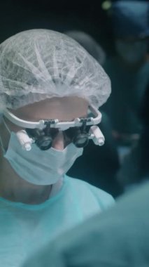 Profesyonel kadın cerrahın dikey göğsü büyüteç içinde asistanlarla konuşurken ameliyathanede ameliyat yapıyor.