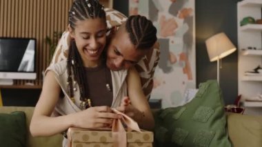 Orta boy mutlu çift ırklı kız hediye paketini açıyor. Sevecen erkek arkadaşı ayakta durup ona sarılıyor ve hediye olarak yeni saç kurutma makinesi alıyor.
