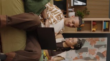 Modern apartmandaki kanepede oturup dizüstü bilgisayarla erkek arkadaşına sataşan ve gıdıklayan komik melez kızın dikey çekimi.