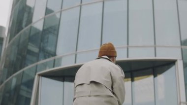 Arka manzara orta yavaş, uzun trençkot giyen bir adam merdivenlerden yukarı, modern cam ofis binasına doğru yürüyor.