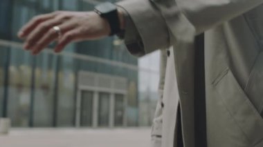 Uzun trençkotlu genç Asyalı bir adamın şehirde karşıya geçerken akıllı saat üzerinden kısa mesaj yazarken görüntüsü.
