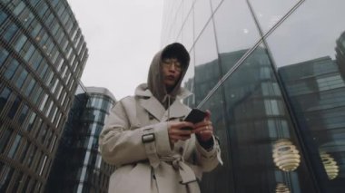 Şehir merkezindeki cam binalar boyunca yürürken Asyalı bir adamın akıllı telefon kullandığı düşük açılı, yavaş çekim.