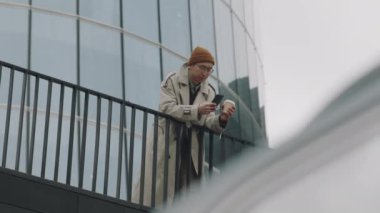 Genç Asyalı bir adamın modern cam duvarlı iş merkezine yaslanıp kahve içerek akıllı telefon kullandığı geniş bir fotokopi.