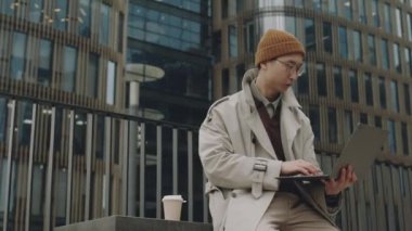 Orta boy Asya 'lı genç bir adam kahve ve dizüstü bilgisayarla bankta oturuyor, modern iş bölgesinde uzaktan çalışıyor.
