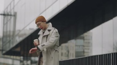 Orta boy Asyalı bir adamın elinde bir fincan kahveyle şehir merkezinde volta atarken saatine bakarken çekilmiş bir fotoğrafı.