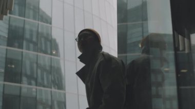 Şehir merkezindeki cam ofis binalarına karşı duran, trençkot giyen Asyalı genç bir adamın orta boy portresi.