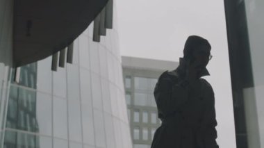 Orta boy, uzun cam ofis binasının önünde yürürken cep telefonuyla konuşan Asyalı genç adamın koyu renk silüeti.