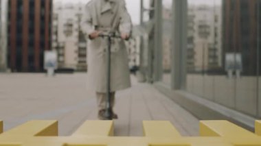 Genç Asyalı bir adam e-scooter kullanıyor ve şehir merkezindeki modern ofis binasının önüne park ediyor.