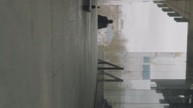 Uzun trençkotlu genç Asyalı adamın gündüz vakti şehirde yürüyüşünün dikey görüntüsü.