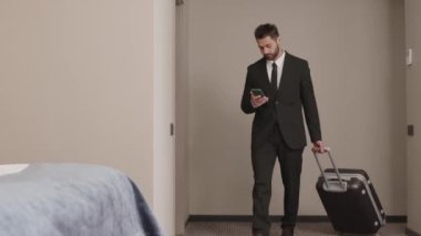 Orta Doğulu genç iş adamı resmi takım elbise giyip elinde bavulla otel odasındaki yumuşak yatağa uzanmış ve gözünü akıllı telefondan ayırmıyor.