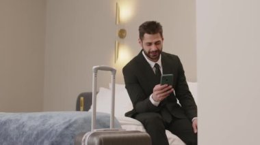 Orta boy sakallı genç Orta Doğulu iş adamı valizinin yanındaki otel odasında yumuşak kral yatağında oturuyor ve cep telefonu kullanıyor.