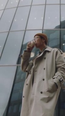 İyi görünümlü Asyalı genç adamın dikey görüntüsü kahve içerken iş bölgesinde uzun cam binalarla dikilirken.