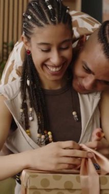 Dikey orta boy mutlu çift ırklı kız hediye paketini açıyor. Sevecen erkek arkadaşı ayakta durup ona sarılıyor ve hediye olarak yeni saç kurutma makinesi alıyor.