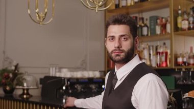 Sakallı, yakışıklı Orta Doğulu barmenin orta boy portresi. Arka planda cam alkol şişeleriyle pahalı barın tezgahında poz veriyor.