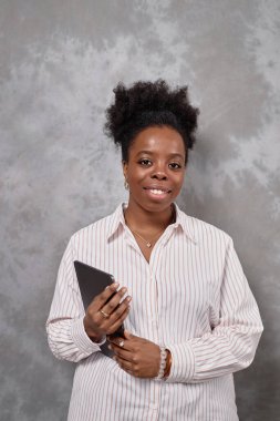 Beyaz tişört giyen, tablet tutan ve gri arka planda izole edilmiş kameraya gülümseyen Afro-Amerikan kadın uzmanının orta boy portresi.