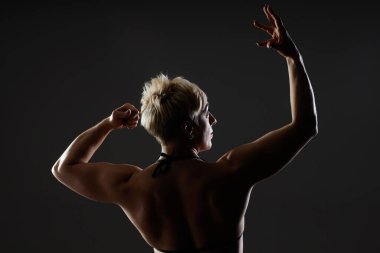 Artistik bir şekilde kollarını kaldırarak poz veren bayan sporcunun orta arka plan çekimi. Vücut geliştiricinin arka plan portresi siyah arka plandaki karanlık stüdyoda sırt kaslarını gösteriyor, boşluğu kopyalıyor