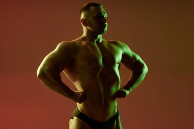 Genç erkek vücut geliştiricinin stüdyo görüntüleri göğüs ve karın kaslarını gevşetiyor. Kırmızı zemin üzerinde yeşil ve nötr ışık olan orta boy atlet görüntüsü, boşluğu kopyala