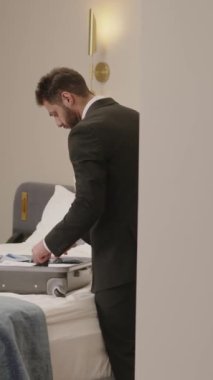 Orta Doğulu resmi takım elbiseli işadamının modern otel odasında kral yatağının yanında duran ve iş gezisine geldiğinde bavulunu boşaltan dikey görüntüsü.