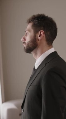 Modern otel odasının penceresinde duran resmi takım elbiseli yakışıklı esmer iş adamının orta boy dikey görüntüsü ve telefon konuşması.