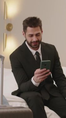 Orta boy sakallı genç Orta Doğulu iş adamının otel odasındaki yumuşak kral yatağında bavulunun yanında otururken ve cep telefonu kullanırken çekilmiş resmi.