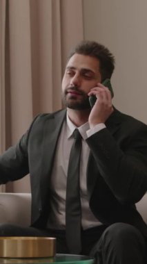 Kendine güvenen Ortadoğulu iş adamının iş gezisi sırasında telefon görüşmesi yaparken, otel odasının penceresinin yanındaki koltukta otururken orta boy dikey çekimi.