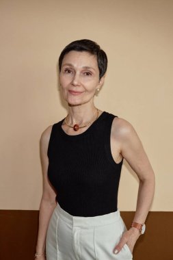 Zarif olgun kadının dikey belden yukarı portresi kameraya gülümseyen nötr renklerle duvarın önünde flaşla çekilen