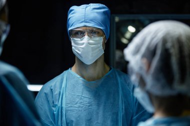 Ameliyat odasında önlük ve koruyucu ekipman giyen erkek cerrahın portresi mavi ışık fotokopi alanı ile aydınlatıldı.