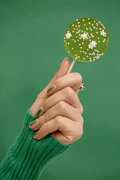 Stock image Vertical studio shot of sugar candy with flowers in female hand. Elegant nude manicure decorated with green streak on emerald background, copy space