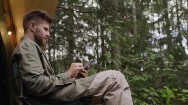 Genç beyaz bir adamın yaz ormanlarında kamp çadırının dışında katlanır sandalyede otururken akıllı telefon kullanırken orta açıdan çekilmiş bir fotoğrafı.