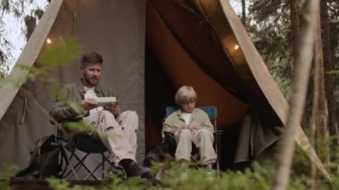Ergen oğlu olan genç, beyaz bir adam ormanda kamp çadırının dışında katlanan sandalyelerde oturuyor, öğle yemeğinde ev yapımı sandviçler yiyor ve sohbet ediyor.
