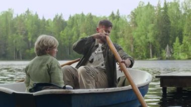 9 yaşındaki oğluyla birlikte balıkçı teknesinde yelken açarken kürek kullanan genç beyaz bir adam.