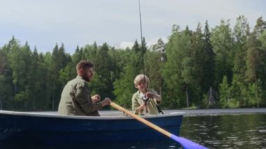 9 yaşında beyaz bir çocuk elinde olta tutarken babasıyla nehirde balık tutarken gün ışığında teknede yan yana oturuyor.
