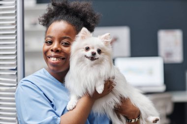 Siyahi etnik kökenli genç bayan veterinerin portre çekimi. Gülümseyen ve kameraya bakan. Genel sağlık kontrolünden sonra mutlu köpek yavrusunu yakın tutarken.