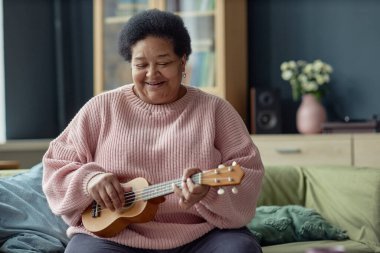 Üst düzey siyahi bir kadının ukulele çalarkenki portresi ve evinde mutlu bir şekilde gülümsemesi. Emeklilik fotokopi odasında yaratıcı hobisinin keyfini çıkarıyor.