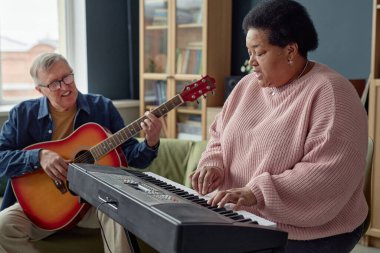 Arka planda gitar çalan son sınıf öğrencisiyle piyano çalan ve şarkı söyleyen siyahi bir kadının yan görünüm portresi