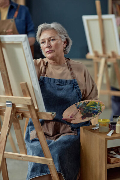 Вертикальный портрет элегантной кавказской женщины, рисующей на мольберте в художественной студии и держащей палитру масляной краской