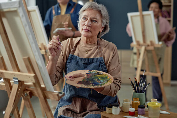 Waist up portrait of creative Caucasian senior woman painting picture on easel and wearing apron while enjoying hobby in art studio copy space