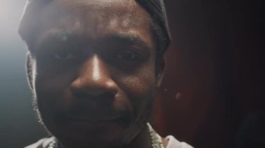 Arkasında projektör ışığı olan siyah arka plandaki kameraya işaret eden ve el kol hareketi yapan, ters şapka takan genç Afrikalı Amerikalı rapçinin belini kaldırın.