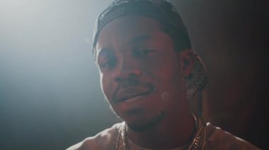 Beyzbol şapkası ters takmış siyah bir adam ve arka planda ışık kaynağıyla kameraya rap yapan birkaç zincir gerdanlık. Yumuşak halo etkisi yaratıyor.